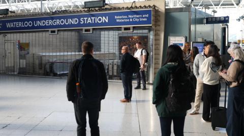 Commuters stand outside the closed entrance to Waterloo Underground Station, where shutters are pulled down.