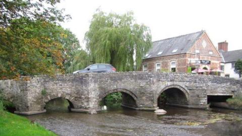 An old-looking stone bridge with a black car driving over it. The bridge is built across a river which passes underneath through five arches. There is some weed on the water. There is a grassy bank to one side of the river, and in the background are two leafy trees. On the other side is an old building, with two tables outside with parasols above them