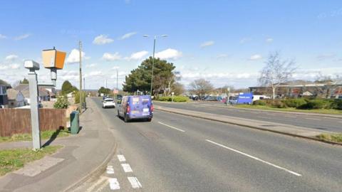 The back of a speed camera can be seen on the left aimed towards a dual carriageway that has a van and car driving on it. A retail park can be seen in the background. 