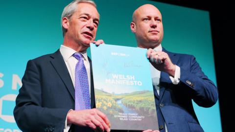 The image shows Nigel Farage and Dan Thomas stood side by side holding a copy of reform UK's Welsh manifesto. They are both wearing navy suits. Farage has a purple tie. They are standing in front of a Reform UK backdrop. 
