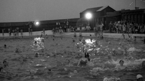 Dozens of people jumping around in an outdoor swimming pool with buildings with floodlights.