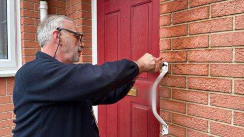 Roger Hewish fitting a white grabrail to the side of a red front door on a brick house. He has white hair and a white beard, wearing glasses and a blue fleece.