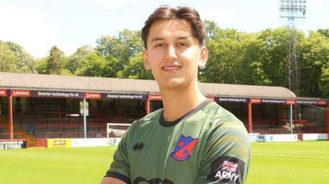 Jack Matton head and shoulders shot smiling at camera in green and black training top in front of club's East Bank terrace
