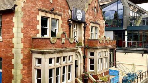 An image of an Edwardian style building with a bridge just visible in the far right hand corner of the image. It has a decking area at the front with seating  