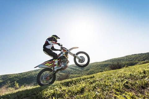 A person on a motocross bike pulling a wheelie. Grass in the foreground and hills in the background against a sunny sky. 