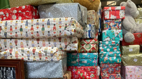 A picture of a number of wrapped presents stacked on top of one another. They are a number of different colours, shapes and sizes. There is a grey bear to the right of the image