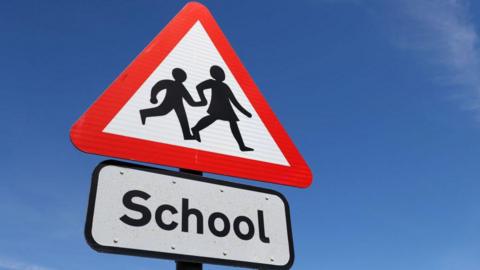 A UK warning triangle road sign, indicating that a school is nearby. The sky is blue.