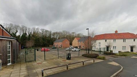 A view of some of the housing on the new estate. There is plenty of parking around a paved square with lots of trees in the background.