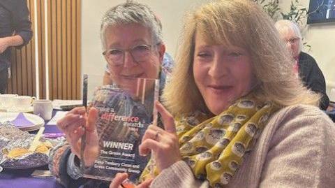 Two women hold a glass Make a Difference award as they sit at a table with a purple tablecloth, food on plates and coffee cups. The woman on the left has white hair and glasses and the woman on the right has blonde hair, a yellow patterned scarf and beige jumper.