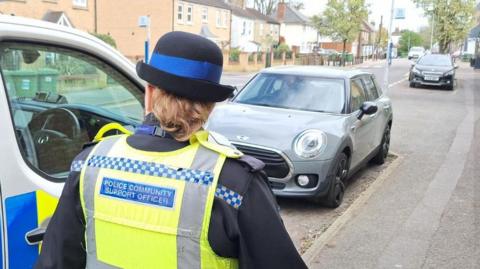 Police support officer in uniform walks along a street from a police vehicle. Cars are parked in laybys.