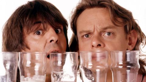 Martin Clunes and Neil Morrissey looking through a row of pint glasses half full of beer.