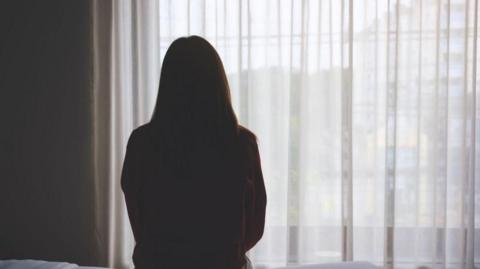 A woman with long dark hair sits in the dark on the edge of a bed facing a window with white net curtains drawn with her back to the camera