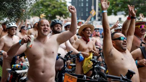 This image as a hundred of half naked people waving their hands while sat on a bicycle. Most of the identifiable people in this picture are men. One man is holding a drink bottle and most of the others are wearing helmets with sun glasses.
