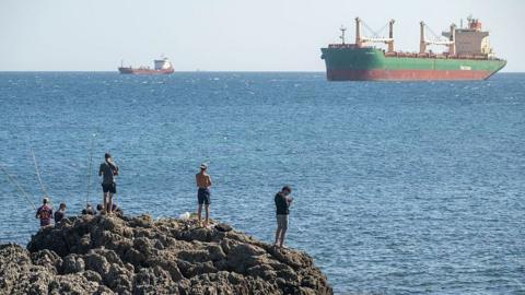 A group of men are seen from the back on a rocky outcrop with the blue sea and large ships in the background, on 22 August, 2026 in Ceuta, Spain.