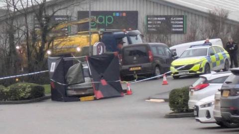 The scene where the car was found. A silver car has been surrounded by black boards and tarpaulin but the back of it is still visible. There is a parked police car and two policemen as well as police scene tape stretched around the car via traffic cones. A metal warehouse-type building is in the background with a sign reading GAP.