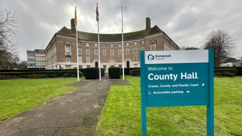 The outside of County Hall in Taunton. There is a pavement and a green lawn in the foreground along with a sign saying "Welcome to County Hall." There is a large building in the background along with three flag poles.