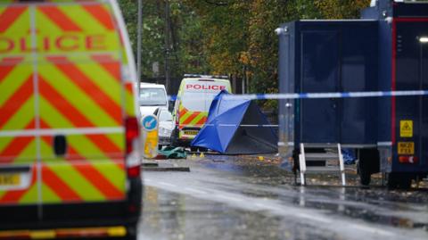 Police van and police cars near the attack scene which has a blue tent in the background