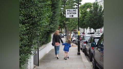 A woman and a young child walking down a tree-lined suburban street hand in hand. Attached to a lamppost is a sign that says 'polling station'.