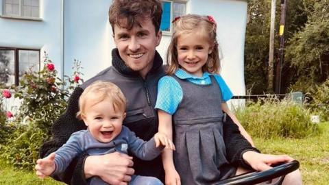 A young dad is sat on a red ride-on lawn mower with his two children in his lap. One is a young blonde boy aged around 16 months, the other is a five-year-old girl with light brown hair in plaits. The girl is wearing her school uniform. 