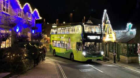 Green bus travelling through road with buildings decorated in lights either side - it is night.