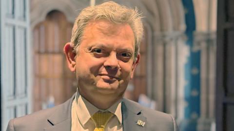 Anton Muscatelli stands inside Glasgow University. The background is the Gothic architecture of the university's main buidling. He is framed by two this wooden doors. Sir Anton wears a grey jacket with a white shirt and yellow tie.