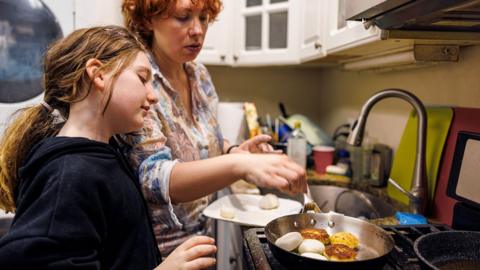 Stock photo shows a mother and daughter standing over a pan cooking on gas in a kitchen