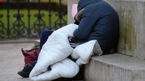 Two rough sleepers sitting at the foot of a stone wall, wrapped in winter coats and hats and with a duvet. They have their heads bowed as if in sleep. There are park railings in the background with a park beyond