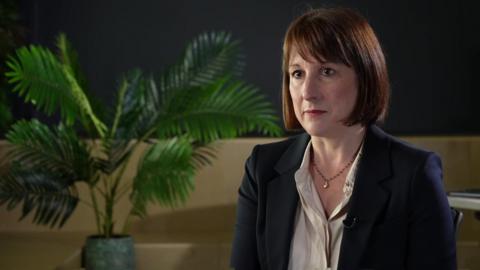 Rachel Reeves sat in front of a fern and a blue wall in a tv interview