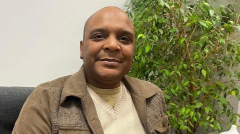 Maurice Blake, who is black, is sitting on a grey couch in front of a plant. He is smiling at the camera. 