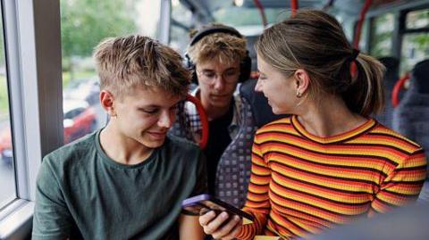 Three children sat on the upper deck of a double-decker bus. A girl wearing a stripey top is holding a phone, showing two other young passengers what's on it.