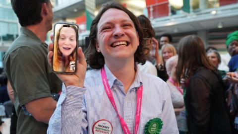 Green Party supporters congratulate candidate Florence Cullen-Davies - who is at home about to give birth and appears live via WhatsApp video on a mobile phone - after she is declared a winner in the 2026 London Borough of Hackne