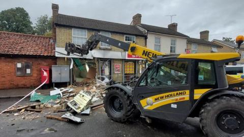 A destroyed shop front with debris on the road and a telehandler