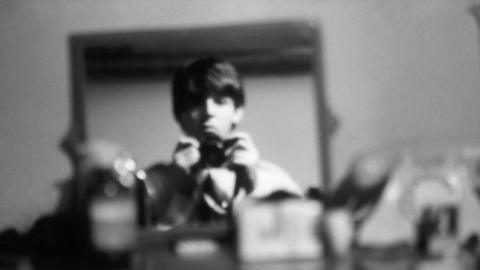 Titled: Self-portrait in my room at the Asher family home, Wimpole Street, London, December 1963. A black and white, out of focus image of Paul McCartney holding his camera in both hands, and pointed at a mirror. He has a straight face and does not smile. 