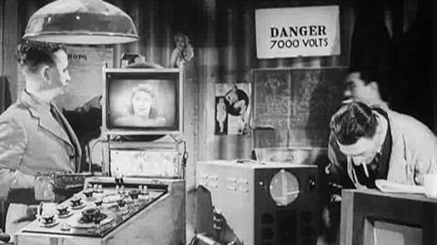 Two radio men stand beside a tv and pictorial equipment