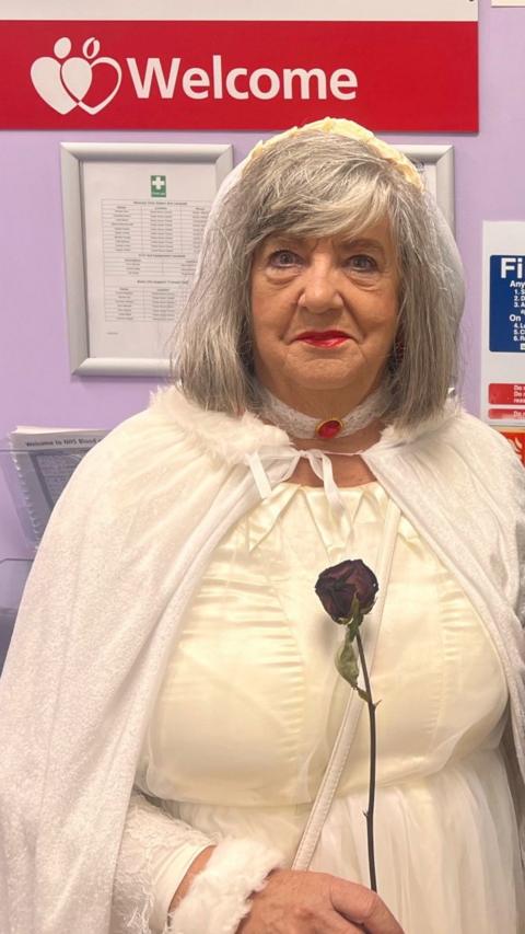 Carol stands in front of a red welcome sign. She has short grey hair and wears a white hooded cloak and a white dress