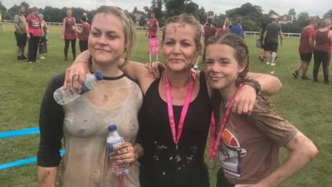 Three women pose for a photo having done a muddy run. The mother is in the middle wearing a black top and medal with a pink lanyard. She has her arms around her two daughters. The one on the right is a teenager, with brown plaited hair and wearing a brown top and a medal. On the left is an older woman in her twenties, wearing a light top and holding a water bottle.