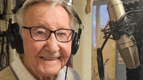 Mr Rayner looking at the camera and smiling. He has brown eyes and white hair. He is wearing headphones and is sitting in front of a microphone.