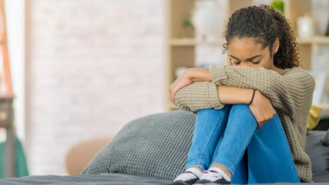 Young girl wearing jeans and a jumper sat on a bed with her head in her hands 