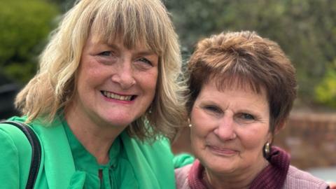 A woman with blonde hair and a green top with a woman who has brown hair and is wearing a pink and purple top