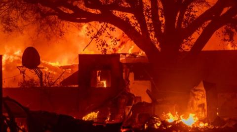 A home is seen burning bright orange with a large tree overhead and a basketball hoop in the background