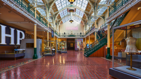 A glass-roofed building with red and blue flooring and items on display in cabinets. There is a green staircase on the right which leads up to the second floor.