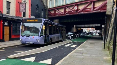 A purple-coloured bus, ahead of other cars, drives past a floating bus stop. There is a small paved section between the main road and the cycle lane, with a pavement on the right hand side. It is situated next to a pub and underneath a red-coloured railway bridge.