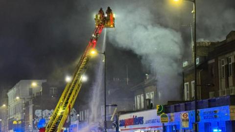 A cherry picker being used by London Fire Brigade containing two firefighters in the top platform spraying water on to the building below. There is a row of shops and the air is smoky. It is also dark as it is early morning.