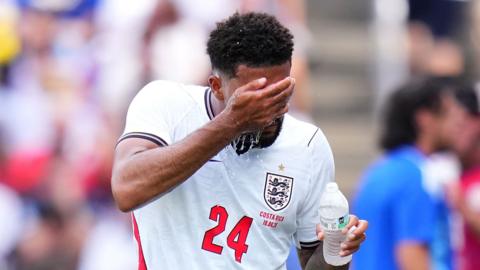 Reece James, wearing an England shirt on the pitch, holding a water bottle and covering his face in water