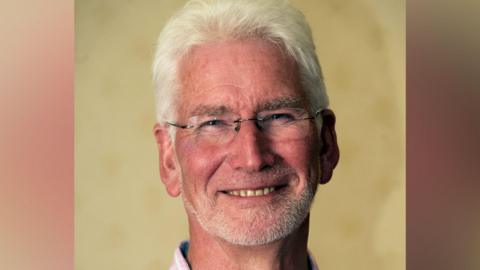 A man, Malcolm Rogers, smiles for a photograph. He has grey hair and a short beard, and wears glasses.