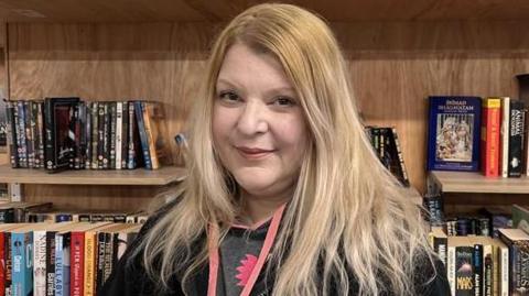 Jo Hawyood smiles at the camera. She has long blonde hair and is wearing a black hooded sweatshirt. She is standing in front of a bookshelf.