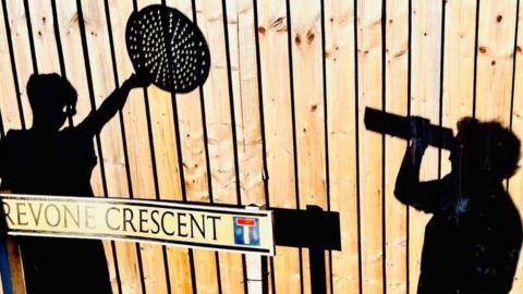 A wooden fence with two shadows cast upon it of people - one holding a cereal box to their eyes and peering through the opening, and the other holding a pizza tray with holes in above their head. There is a street sign at the bottom right of the image with the name 'Trevone Crescent' written on it.