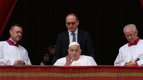 Pope Francis sitting on a balcony waving at a crowd flanked by at least three aides.