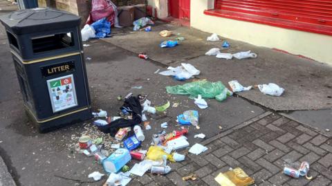 Food waste and litter strewn all over a street in front of the Clarence Hardware & DIY Centre store. The items are right next to a black  council bin.