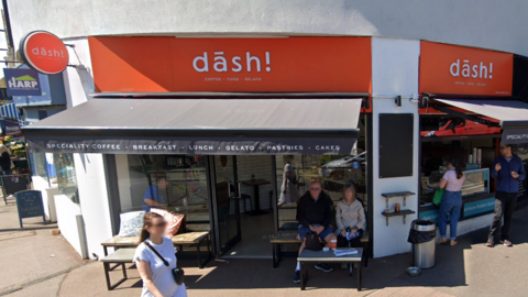 A white building with bright orange sign above the entrance reading "Dāsh!" and black awning reading "Speciality coffee. Breakfast. Lunch. Gelato. Pastries. Cakes". There are two people sitting at tables in front of the shop and two more waiting at the side of the building at the ice-cream window.